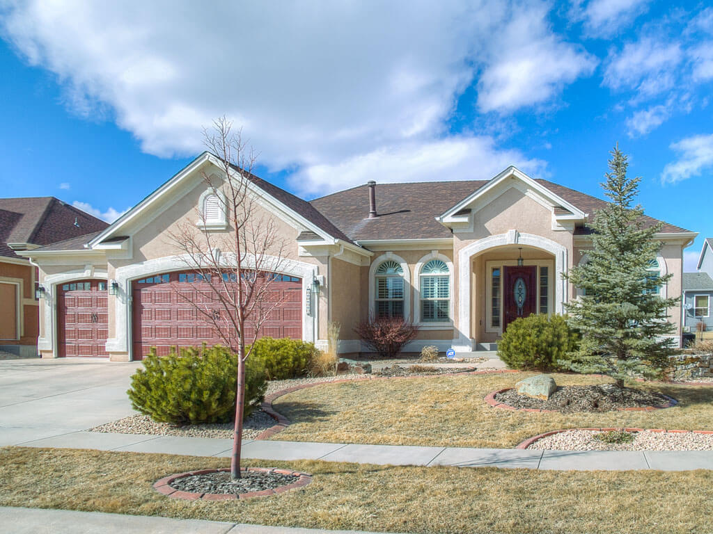 Single-story suburban home with beige stucco exterior, three-car garage with dark red doors, arched windows, red front door, and drought-tolerant landscaping with shrubs, rocks, and a bare tree under a bright blue sky with scattered clouds